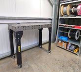 A heavy duty modular welding table with black legs and a gray tabletop, on castor wheels, set in a workshop environment.